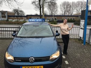 Een glimlachende vrouw staat naast een blauwe Volkswagen rijschoolauto met een RIJ-WIJS rijschoolbord op het dak en maakt een vredesteken op een parkeerplaats met gebouwen en bladerloze bomen op de achtergrond.