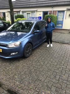 Een persoon in een blauwe hoodie staat op een natte straat naast een blauwe rijschoolauto met een DRIJWIS-rijschoolbord op het dak, geeft een duim omhoog en glimlacht voor huizen met gesnoeide struiken.