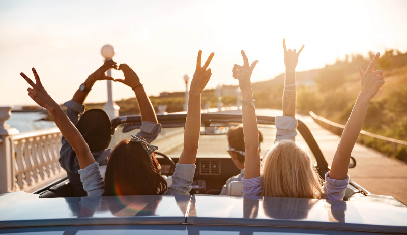 Drie vrienden in een cabriolet van een rijschool met het dak naar beneden, rijdend langs een zonnige kustweg, hun armen omhoog en maken vrolijke handgebaren, feestend en genietend van het moment.